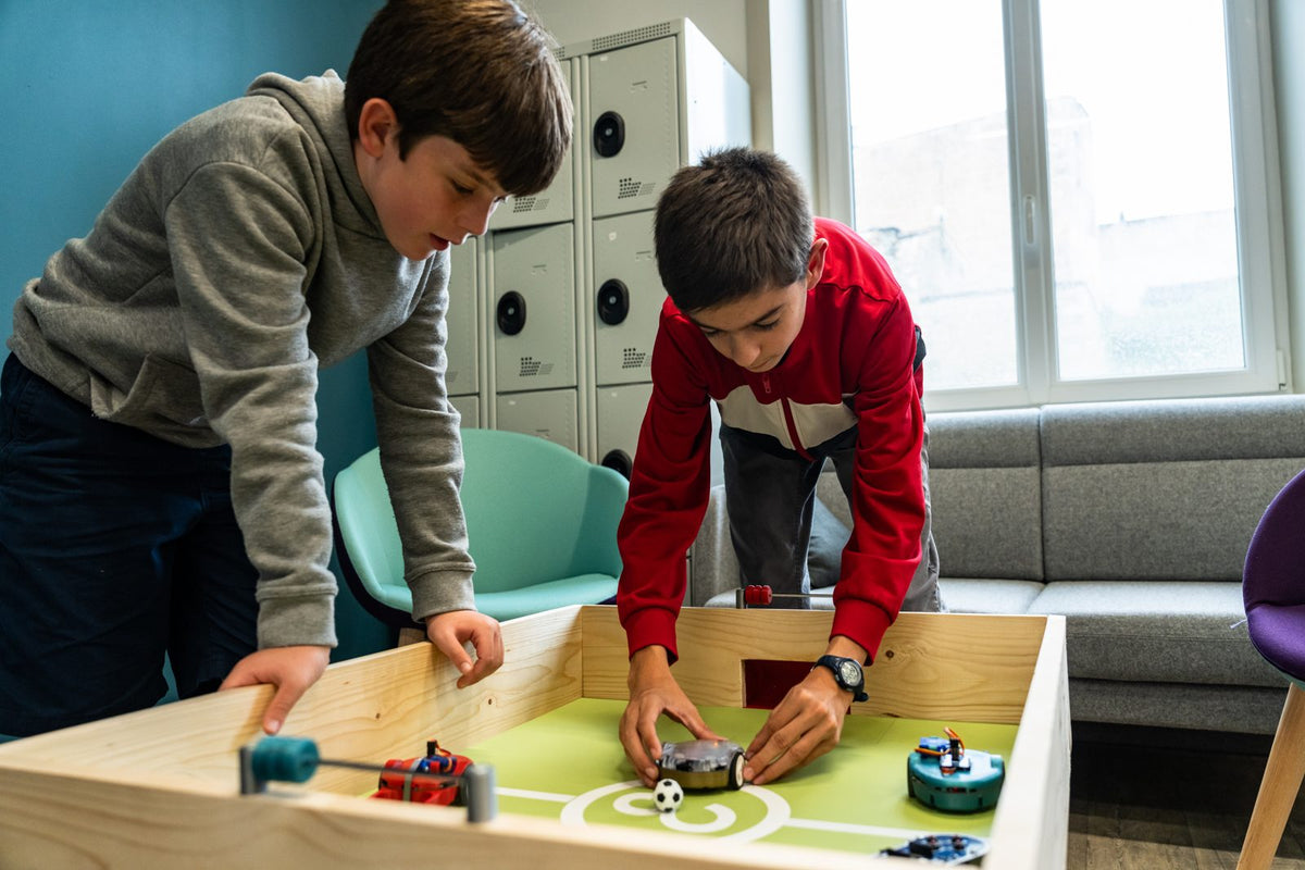 Deux enfants organisent un match de foot entre robots Eliobot sur un terrain en bois
